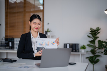 Young professional woman in a suit engaging with a laptop, presenting financial charts and graphs while explaining data during a video conference in a modern office setting