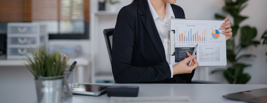Businesswoman sitting at office desk pointing at financial charts and graphs using a pen, analyzing company performance and making strategic decisions based on market data