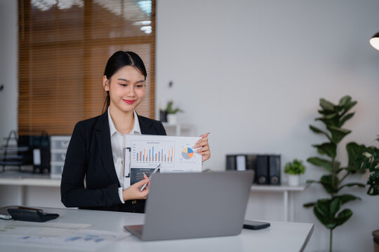 Young professional woman in a suit engaging with a laptop, presenting financial charts and graphs while explaining data during a video conference in a modern office setting