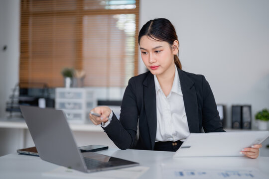 Young Asian businesswoman holding a pen while analyzing sales data on paperwork, working diligently on a laptop at her modern office desk, surrounded by bright natural light