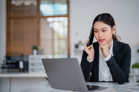 Young businesswoman is sitting at her desk, talking on her phone and working on her laptop, concentrating on her tasks in a bright and modern office environment