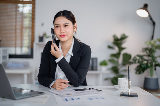 Asian businesswoman dressed in a sleek black suit, talking on a mobile phone while analyzing financial charts and working diligently on a laptop at her office desk - Powered by Adobe