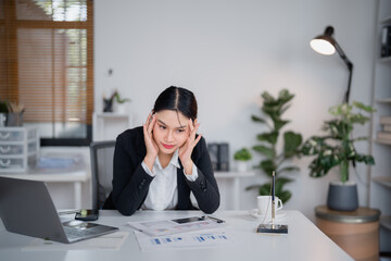 Young Asian businesswoman is having a headache and touching her temples while working with laptop...