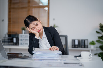 Asian businesswoman resting her head on her hand while working on a large pile of documents on her desk in the office, feeling tired and stressed