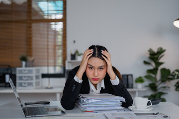 Businesswoman sitting at desk, holding her head in her hands from overwhelming stress, surrounded...