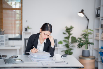 Young asian businesswoman having headache and taking notes on documents at office desk, she is...