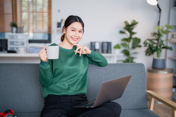 Beautiful smiling asian freelancer woman drinking coffee and working on laptop sitting on sofa at home, she is relaxing on comfortable sofa in living room
