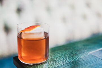 Elegant cocktail with ice cube and orange peel garnish in a clear glass on a green wooden surface,...