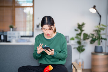 Young woman sitting on a sofa in a cozy living room, struggling with her smartphone while...