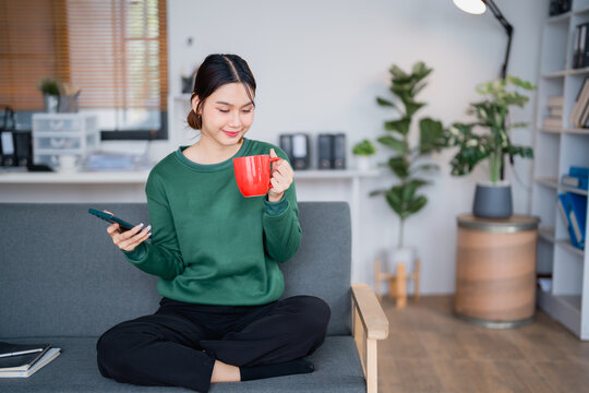 Freelancer woman is enjoying a cup of coffee while checking her smartphone, sitting comfortably on the sofa in her home office, embracing the flexibility of remote work
