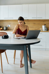 Young female freelancer in pajamas keeping track of her day sitting at table with coffee and laptop