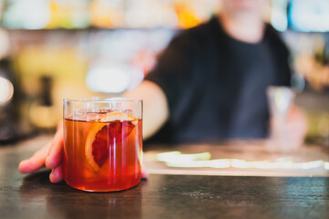 Bartender serving a cocktail with orange garnish in a clear glass on bar counter, blurred background, focus on hand and drink, concept of nightlife, hospitality, and mixology.