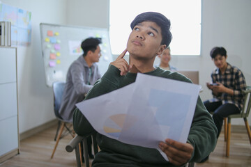 Young Asian Man Deep In Thought Holding Chart And Looking Upwards