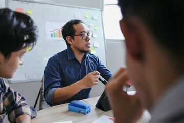 Asian Male Teacher Wearing Glasses Speaks To Students Around Table With Digital Tablet