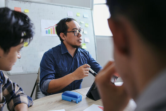 Asian Male Teacher Wearing Glasses Speaks To Students Around Table With Digital Tablet - Powered by Adobe