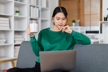 A cute Asian woman is working remotely from a home office, enjoying coffee while working on laptop 