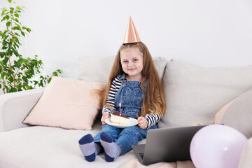 Birthday celebration. Little girl with piece of cake having video call via laptop on sofa at home