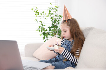 Birthday celebration. Little girl blowing candle on piece of cake while having video call via...