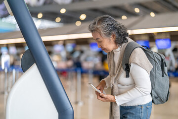 Asian senior woman traveler passenger with luggage using digital check in at airport kiosk, smart...