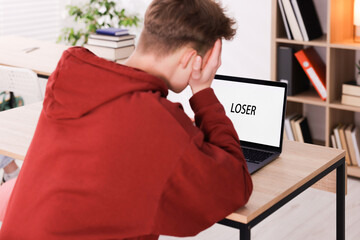 Cyber bullying. Upset teenage boy and laptop with text You Loser at desk indoors