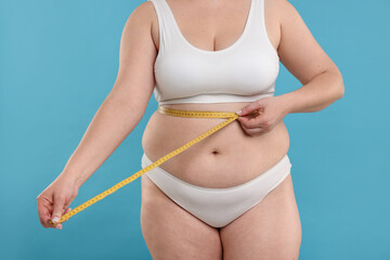 Weight loss. Plus size woman measuring waist with tape on light blue background, closeup