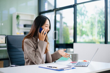 Confident young Asian businesswoman smiles while multitasking talking on her smartphone and using her laptop at a modern office desk