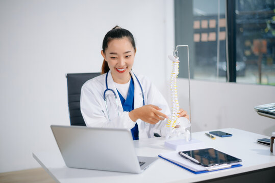 young Asian doctor holds a spine model and uses a tablet at her desk showcasing real people, medical expertise, and modern healthcare education - Powered by Adobe