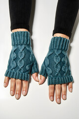 A close-up of hands resting on a white surface, wearing fingerless cable knit gloves in shades of teal and blue. The gloves feature a thick, ribbed cuff at the wrist.