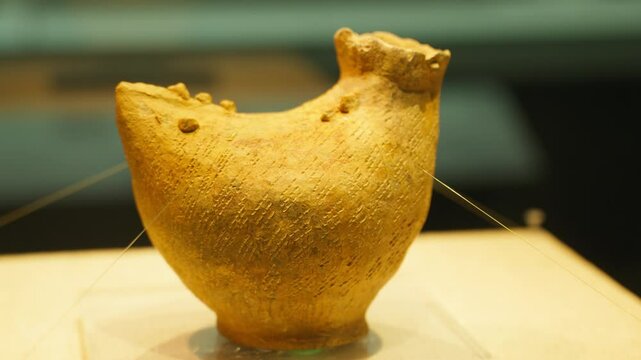 A rugged yellow-brown ceramic vessel with asymmetrical shape and display strings sits in a museum setting&mdash;highlighting ethnic craftsmanship, cultural depth, and the tactile presence of historical pott