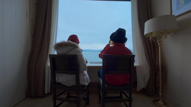 Two people in winter attire sit quietly by a large window, gazing out at a serene waterscape&mdash;featuring soft indoor lighting, travel jackets, and the tranquil rhythm of alpine rest and contemplation.