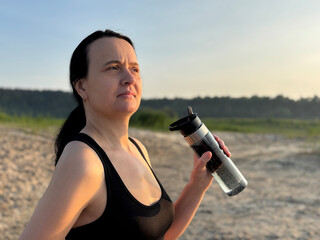 Close-up of athletic woman hand holding shaker bottle with water on sandy trail near forest,...