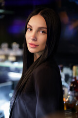 Fototapeta premium Confident young woman with long black hair and natural makeup posing indoors in elegant black outfit, standing near a bar with soft evening lights in the background. Fashion and lifestyle concept.