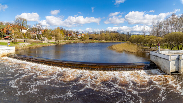 Pskov Dam on Pskova River with turbulent water flow over concrete spillway, surrounded by natural grassy banks and spring greenery. Sunny day in Pskov, Russia. 