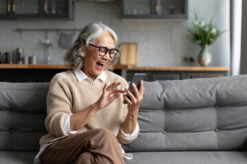 Happy senior woman using smartphone on sofa at home, smiling and laughing while browsing or chatting.