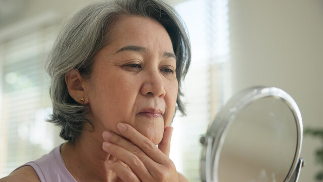 Asia adult people middle age old woman look at mirror dry skin face hand touch neck cheek stress sad at home elder health care aging beauty issue. Asian female aged eye botox filler plastic surgery.