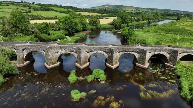 Clady Bridge, County Tyrone, Northern Ireland, June 2023. Drone slow pullback from arched bridge connecting across River Finn, ascending above to establish the countryside.