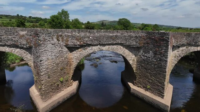 Clady Bridge, County Tyrone, Northern Ireland, June 2023. Drone ascend along symmetrical arch to top shot above as car crosses River Finn.