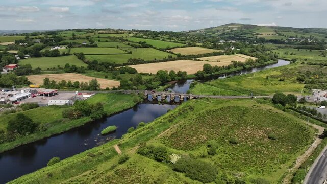 Clady Bridge, County Tyrone, Northern Ireland, June 2023. Drone panoramic orbit ascend clockwise showcasing arched bridge crossing River Finn with clouds reflecting in water and both villages visible.