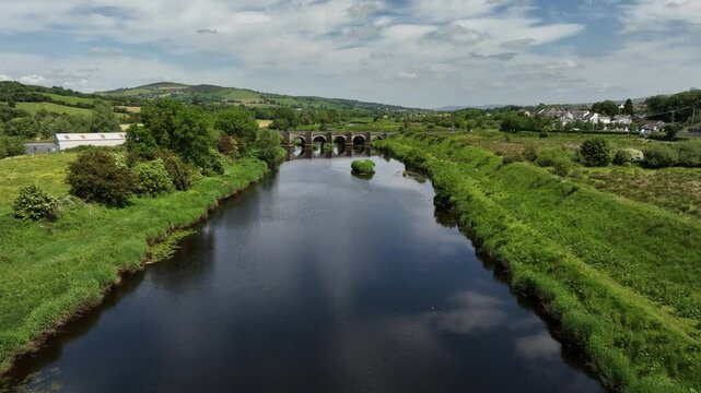 Clady Bridge, County Tyrone, Northern Ireland, June 2023. Drone approach along River Finn to arched bridge with sloped hills in backdrop along grassy riverbanks.