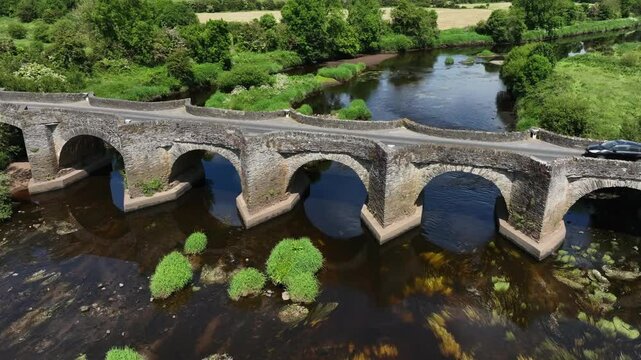 Clady Bridge, County Tyrone, Northern Ireland, June 2023. Drone orbit counterclockwise around arched bridge crossing River Finn as cars passover top on sunny day.