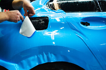 Detailing technician trims blue PPF on a car panel for perfect contour fit. Cutting blue PPF with...