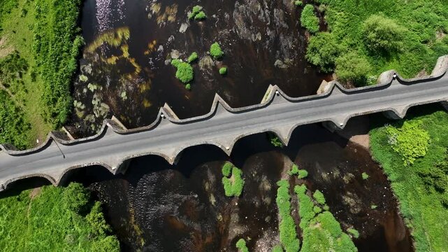 Clady Bridge, County Tyrone, Northern Ireland, June 2023. Drone high bird's eye overview orbit clockwise above arched bridge with water streaming down across rocks in River Finn.