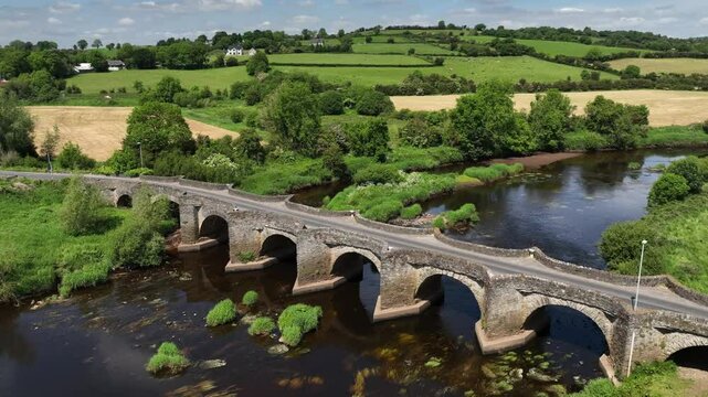 Clady Bridge, County Tyrone, Northern Ireland, June 2023. Drone orbit clockwise descend and zoom in to arched bridge with symmetrical reflection of bricks and clouds in water of River Finn.