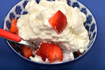 A close-up shows a spoon taking a serving of strawberries and whipped cream from a blue and white patterned bowl against a blue background