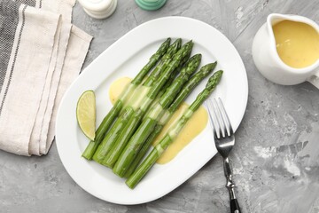 Tasty cooked asparagus served with sauce and lime on grey textured table, flat lay