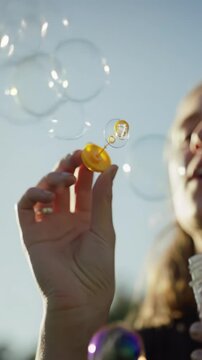 Human hand blows iridescent soap bubbles, macro video capturing vibrant rainbow colors. Shimmering spheres float against a soft, sun-drenched background with bokeh. Slow-motion low-angle video in