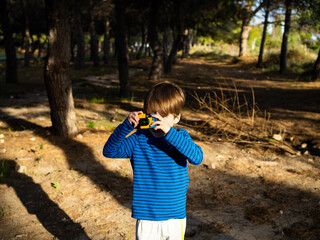 A boy is in a forest focused on capturing photographs with a disposble camera, wearing a blue...