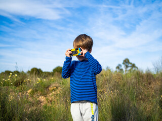 A young boy stands in a grassy field, holding a disposable camera to his eye. The sun shines...