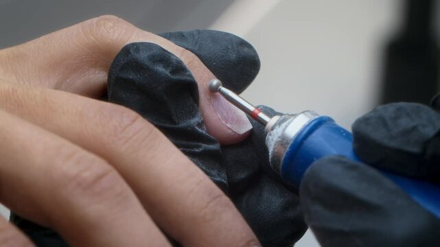 Close-up of gloved technician operating electric manicure tool while working on client nails and cuticles during session in salon
