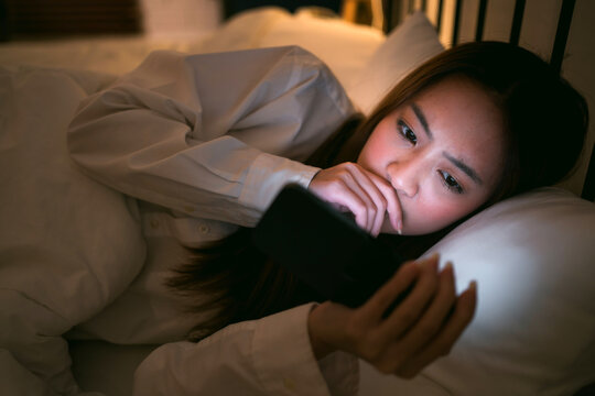 Young woman lying in bed late at night, staring at smartphone screen with concerned expression. Concept of sleep disruption, eye strain, and mental health impact from late-night mobile device use. - Powered by Adobe